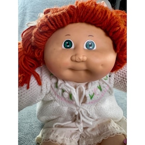 Könnte beinhalten: Eine Puppe mit leuchtend orangefarbenem Wollhaar, das zu Zöpfen mit weißen Bändern gebunden ist. Die Puppe hat ein rundes Vinylgesicht, grüne Augen und ein Grübchen an der linken Wange. Sie trägt eine weiße Strickjacke mit grün-rosa Blumenstickerei.