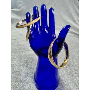 May include: A cobalt blue glass hand sculpture displaying three metal bangle bracelets. Two bracelets are positioned on the fingers, one featuring a smooth beige band and the other a metallic finish. A third gold-toned hinged bangle rests on the thumb.