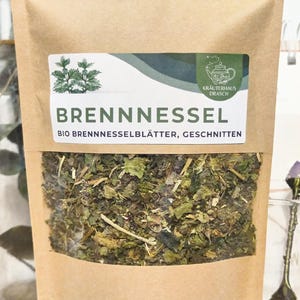 Könnte beinhalten: Ein brauner Papierbeutel mit einem Sichtfenster, in dem getrocknete grüne Brennnesselblätter zu sehen sind. Der Beutel hat ein Etikett mit der Aufschrift "BRENNNESSEL" und "BIO BRENNNESSELBLÄTTER, GESCHNITTEN" in schwarzer Schrift. Das Etikett zeigt außerdem eine kleine Illustration einer Teetasse und einer Pflanze.