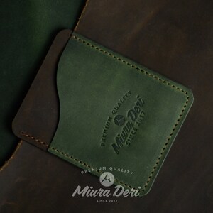Puede incluir: Un porta tarjetas de cuero verde con un acento de cuero marr&oacute;n. El porta tarjetas tiene un borde cosido y el texto "Premium Quality Miura Deri Since 2017" grabado en la parte delantera.