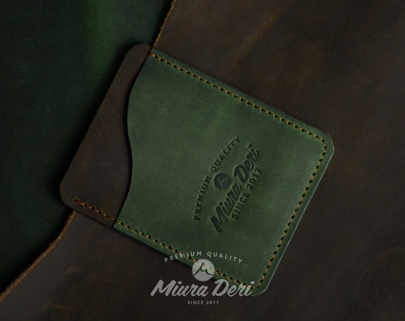 Puede incluir: Un porta tarjetas de cuero verde con un acento de cuero marr&oacute;n. El porta tarjetas tiene un borde cosido y el texto "Premium Quality Miura Deri Since 2017" grabado en la parte delantera.