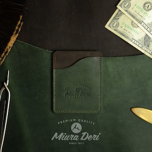 Puede incluir: Un porta tarjetas de cuero verde con una solapa de cuero marr&oacute;n y un borde cosido. El porta tarjetas tiene el texto "PREMIUM QUALITY Miura Deri SINCE 2017" grabado en la parte delantera.