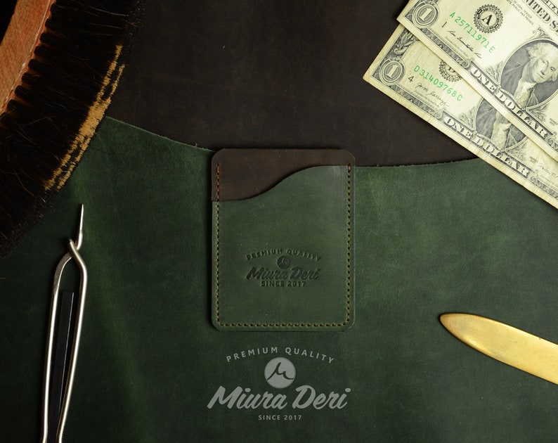 Puede incluir: Un porta tarjetas de cuero verde con una solapa de cuero marr&oacute;n y un borde cosido. El porta tarjetas tiene el texto "PREMIUM QUALITY Miura Deri SINCE 2017" grabado en la parte delantera.