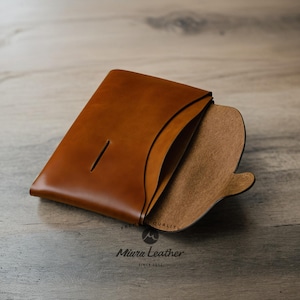 K&ouml;nnte beinhalten: Eine braune Ledergeldb&ouml;rse mit einer Klappe, die sich &ouml;ffnet und ein braunes Wildleder-Innenfutter zeigt. Die Geldb&ouml;rse hat einen einzigen Schlitz auf der Vorderseite und der Text "Miura Leather" ist unten aufgedruckt.