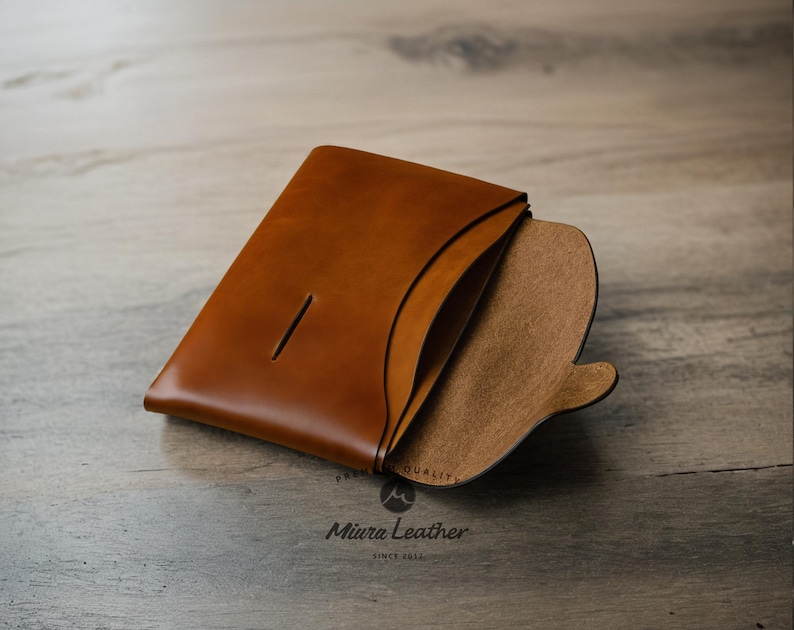 K&ouml;nnte beinhalten: Eine braune Ledergeldb&ouml;rse mit einer Klappe, die sich &ouml;ffnet und ein braunes Wildleder-Innenfutter zeigt. Die Geldb&ouml;rse hat einen einzigen Schlitz auf der Vorderseite und der Text "Miura Leather" ist unten aufgedruckt.