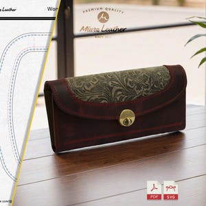 Könnte beinhalten: Eine bordeauxfarbene Ledergeldbörse mit floral gemusterter Klappe und goldfarbenem Verschluss. Die Geldbörse ist rechteckig und liegt auf einer Holzoberfläche. Der Markenname "Miura Leather" ist im Hintergrund sichtbar.