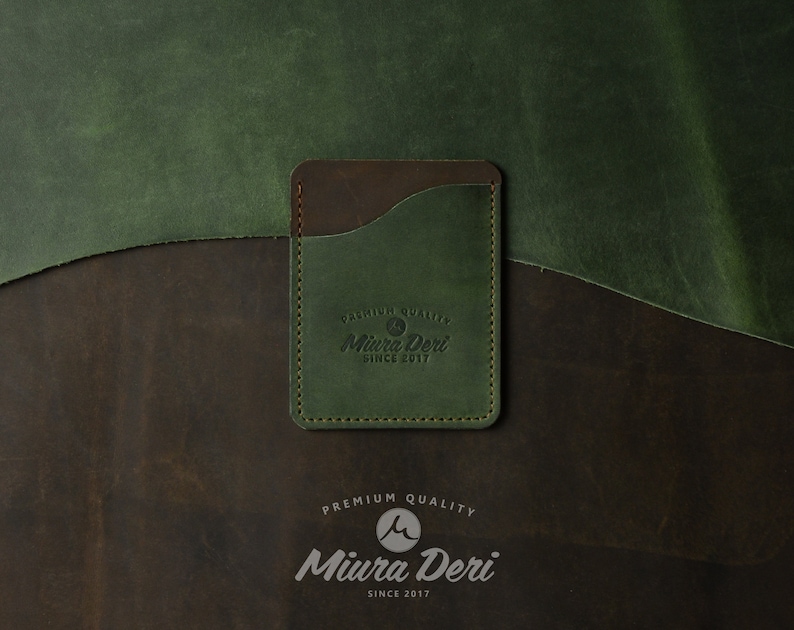 Puede incluir: Un porta tarjetas de cuero verde con una solapa de cuero marr&oacute;n y un logotipo cosido que dice "Premium Quality Miura Deri Since 2017".
