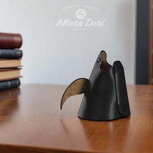 May include: A black leather desk organizer shaped like a stylized bird, with a curved wing and a small eye detail. The organizer sits on a wooden surface, with a stack of books in the background. The Miura Deri logo is visible.