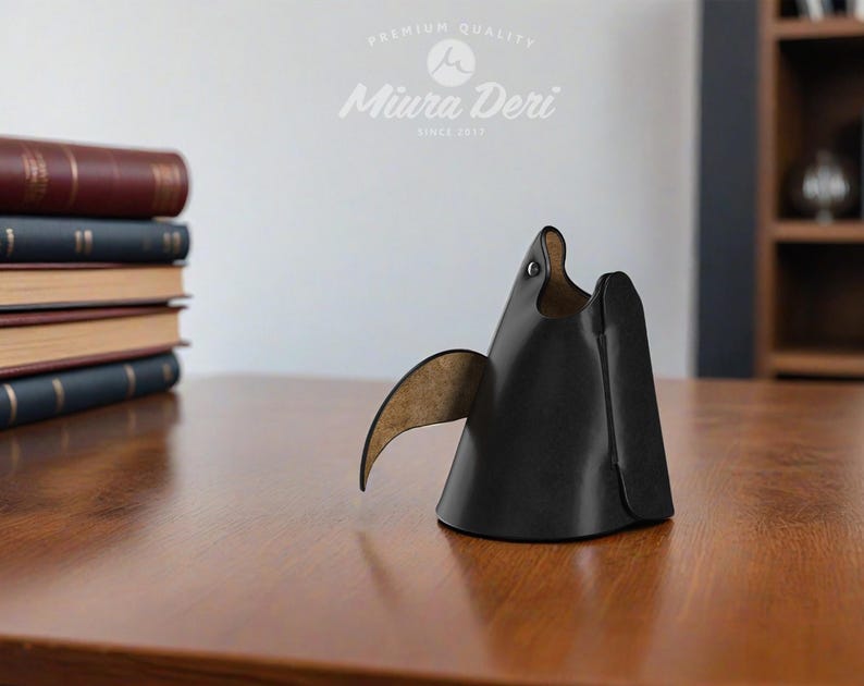 May include: A black leather desk organizer shaped like a stylized bird, with a curved wing and a small eye detail. The organizer sits on a wooden surface, with a stack of books in the background. The Miura Deri logo is visible.