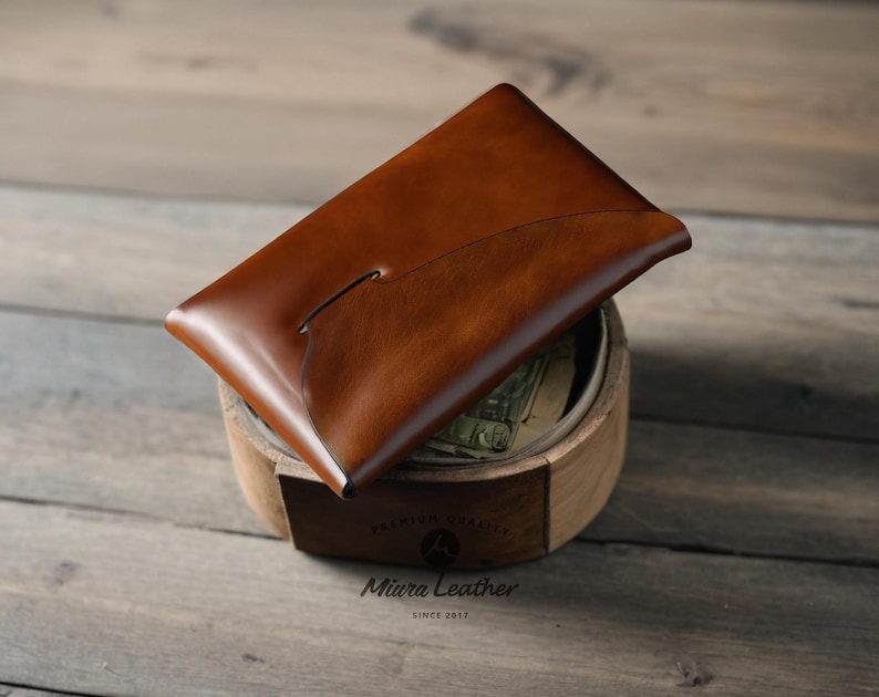 K&ouml;nnte beinhalten: Eine braune Ledergeldb&ouml;rse mit einem einzigartigen, geschwungenen Design. Die Geldb&ouml;rse ist ge&ouml;ffnet und liegt auf einer Holzoberfl&auml;che. Der Text "Miura Leather Since 2013" ist auf der Oberfl&auml;che sichtbar.