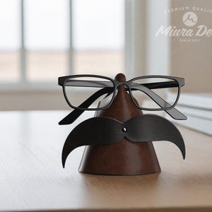 May include: A brown leather glasses and mustache holder with black-framed glasses and a black mustache cutout. The holder is cone-shaped and sits on a wooden surface. The Miura Deri logo is visible in the background.