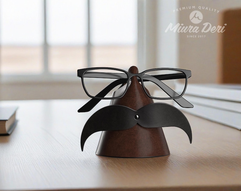 May include: A brown leather glasses and mustache holder with black-framed glasses and a black mustache cutout. The holder is cone-shaped and sits on a wooden surface. The Miura Deri logo is visible in the background.