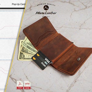 Puede incluir: Cartera de cuero marrón abierta con efectivo y una tarjeta de crédito. La cartera está abierta, revelando varios bolsillos y un cierre de botón a presión. La tarjeta de crédito es negra con texto blanco.