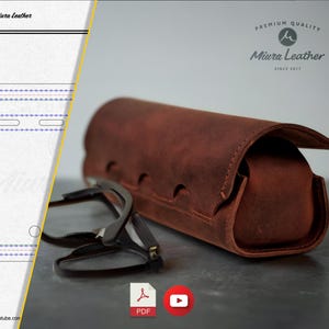 May include: A brown leather glasses case with a flap closure. The case is made of premium quality leather and has a stitched design. The case is sitting on a gray surface next to a pair of glasses.