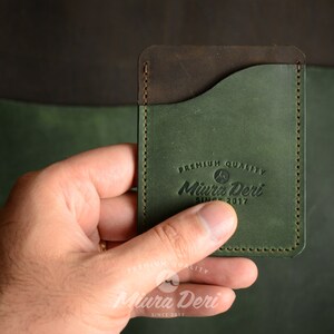Puede incluir: Un porta tarjetas de cuero verde con una solapa de cuero marr&oacute;n y un borde cosido. El porta tarjetas tiene el texto "Premium Quality Miura Deri Since 2017" grabado en la parte delantera.