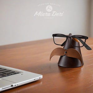 May include: A brown leather glasses and mustache holder with black-framed glasses and a light brown mustache. The holder is cone-shaped and sits on a wooden surface. The text "Miura Deri" is visible.