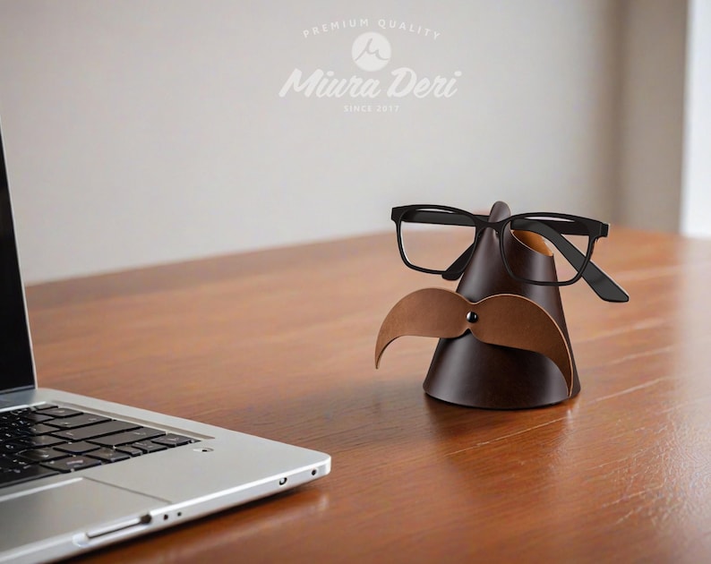 May include: A brown leather glasses and mustache holder with black-framed glasses and a light brown mustache. The holder is cone-shaped and sits on a wooden surface. The text "Miura Deri" is visible.