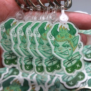 May include: A set of six green and white keychains featuring a cartoon character with green hair, horns, and a mischievous expression. Each keychain has a different design and is attached to a silver key ring.