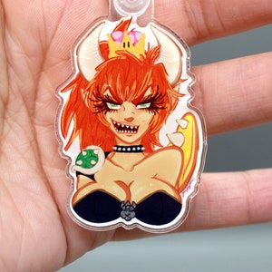 May include: A clear acrylic keychain featuring a cartoon illustration of a female character with red hair, a crown, and a green shell. The character has a fierce expression and is wearing a black outfit.