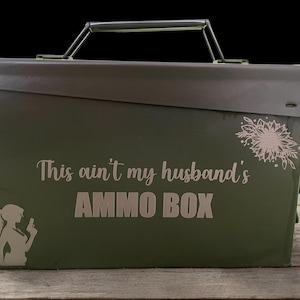 Könnte beinhalten: Eine olivgrüne Munitionskiste mit schwarzem Griff und dem Text "This ain't my husband's AMMO BOX." Ein Blumendesign und eine Silhouette einer Frau mit einer Waffe sind ebenfalls abgebildet.