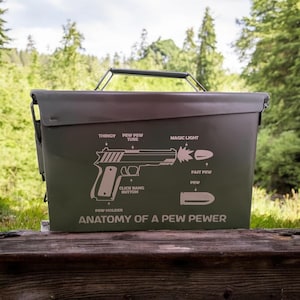Könnte beinhalten: Eine olivgrüne Munitionskiste aus Metall mit einem schwarzen Griff. Die Kiste zeigt ein Diagramm einer Handfeuerwaffe mit Bezeichnungen wie "THINGY" und "MAGIC LIGHT". Der Text "ANATOMY OF A PEW PEWER" ist darunter gedruckt.