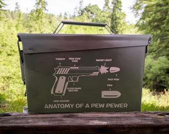 Puede incluir: Una caja de municiones de metal verde oliva con un asa negra. La caja presenta un diagrama de una pistola con etiquetas como "THINGY" y "MAGIC LIGHT". El texto "ANATOMY OF A PEW PEWER" está impreso debajo.