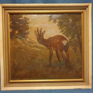 May include: Oil painting of a deer in a forest setting. The deer is brown with a white patch on its rear. The painting has a gold frame with a cream-colored border. The background features trees and a soft, natural light.