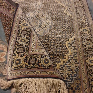 Puede incluir: Una alfombra ornamental enrollada con un detallado patrón floral en tonos azul marino, beige y dorado. La alfombra presenta un borde decorativo y largas borlas beige. El diseño es intrincado y tradicional, adecuado para la decoración del hogar.