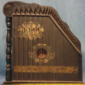 Può includere: Antica cetra nera con corde, con opere d'arte floreali e paesaggistiche decorative. Lo strumento ha una finitura scura con accenti dorati e un piolo di accordatura in legno. Un metro a nastro è in basso.