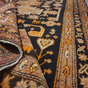May include: A vintage rug with a dark blue field and intricate patterns in shades of orange, brown, and cream. The rug features geometric and floral motifs, with a rolled-up section revealing the underside.