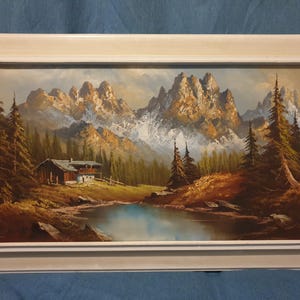 May include: Oil painting depicting a mountain scene with a cabin, lake, and fir trees. The snow-covered mountains are in the background, under a cloudy sky. The artwork is presented in a white rectangular frame.