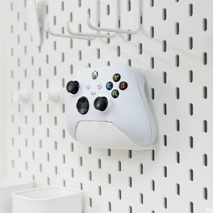 Könnte beinhalten: Ein weißer Xbox-Controller, der an einer weißen Lochwand befestigt ist. Der Controller hat schwarze Joysticks und Tasten mit farbigen Aktionstasten. Zusätzliches weißes Zubehör ist ebenfalls an der Lochwand befestigt.
