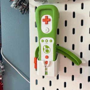 Peut inclure: Une manette Wii verte et blanche avec un pavé directionnel en forme de croix rouge et des boutons orange. La manette est dans un support blanc avec une dragonne verte attachée. La manette est montée sur une plaque perforée blanche.