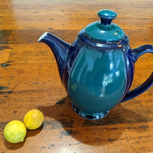 Puede incluir: Una tetera de cerámica azul y verde con tapa sobre una superficie de madera. Dos limones amarillos están en primer plano.