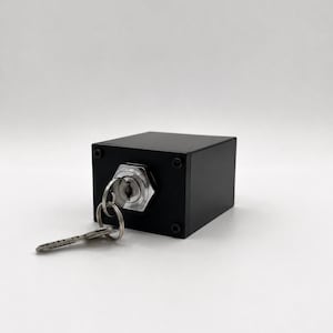 May include: A matte black rectangular box featuring a silver metal key switch mechanism on the front face. A silver metal key is inserted into the circular keyhole, with a small metal keyring attached to the key head, resting on the surface.