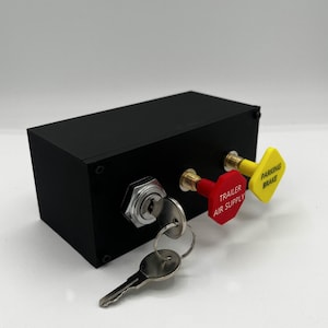 Könnte beinhalten: Ein schwarzer rechteckiger Steuerkasten mit einem Schlüsselschloss und zwei Zugknöpfen. Einer ist rot und mit "TRAILER AIR SUPPLY" beschriftet, der andere ist gelb und mit "PARKING BRAKE" beschriftet. Ein Satz Schlüssel steckt im Schloss.