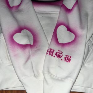 May include: White hoodie with a pink Hello Kitty graphic on the front and pink hearts on the sleeves. The text "M.S.B" is printed on the right sleeve.