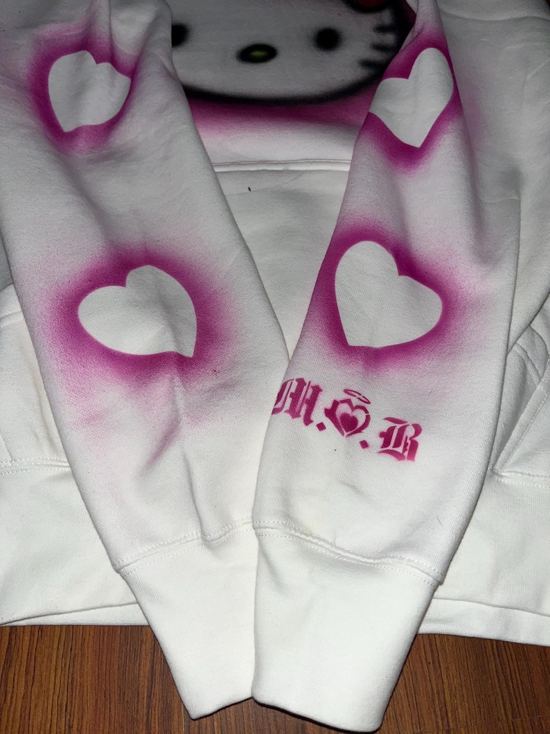 May include: White hoodie with a pink Hello Kitty graphic on the front and pink hearts on the sleeves. The text "M.S.B" is printed on the right sleeve.
