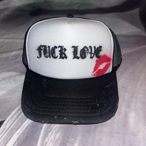Puede incluir: Gorra de camionero en blanco y negro con el texto "FUCK LOVE" en fuente gótica negra. Una marca de beso de lápiz labial rojo está en el lado derecho. La gorra tiene una visera negra y un panel frontal blanco.