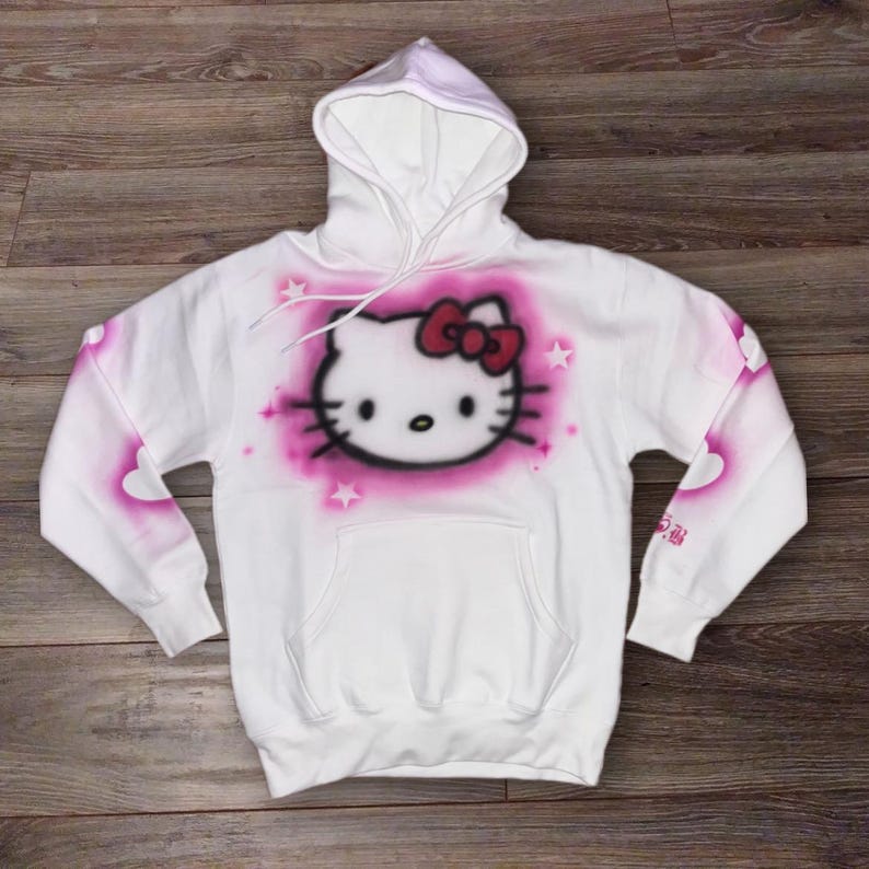 May include: White hoodie with a pink and white Hello Kitty graphic on the front. The graphic is surrounded by pink stars and hearts. The hoodie has a hood and a kangaroo pocket.