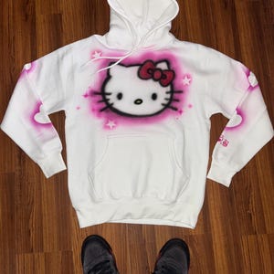 May include: White hoodie with a pink Hello Kitty graphic spray painted on the front. The hoodie has a pink heart design on the sleeves.