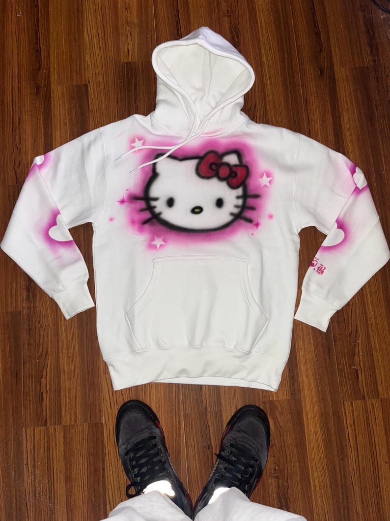 May include: White hoodie with a pink Hello Kitty graphic spray painted on the front. The hoodie has a pink heart design on the sleeves.