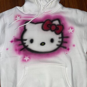 May include: White hoodie with a pink and red Hello Kitty graphic spray painted on the front. The graphic includes a pink bow and white stars.