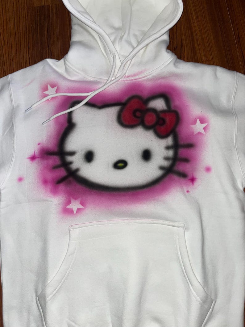 May include: White hoodie with a pink and red Hello Kitty graphic spray painted on the front. The graphic includes a pink bow and white stars.