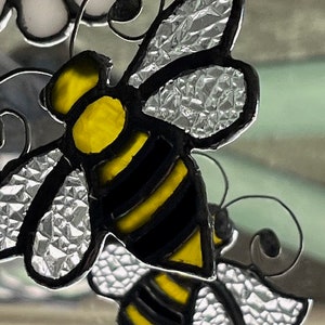 May include: A stained glass bee with black and yellow stripes and clear wings. The bee is hanging from a wire and is part of a larger stained glass design.