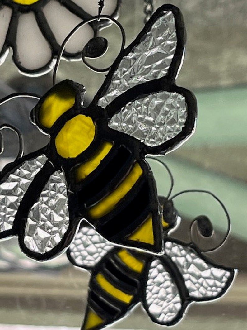 May include: A stained glass bee with black and yellow stripes and clear wings. The bee is hanging from a wire and is part of a larger stained glass design.