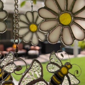 May include: A stained glass wind chime featuring white daisies with yellow centres and clear glass bumblebees with black stripes. The wind chime is hanging from a silver chain.