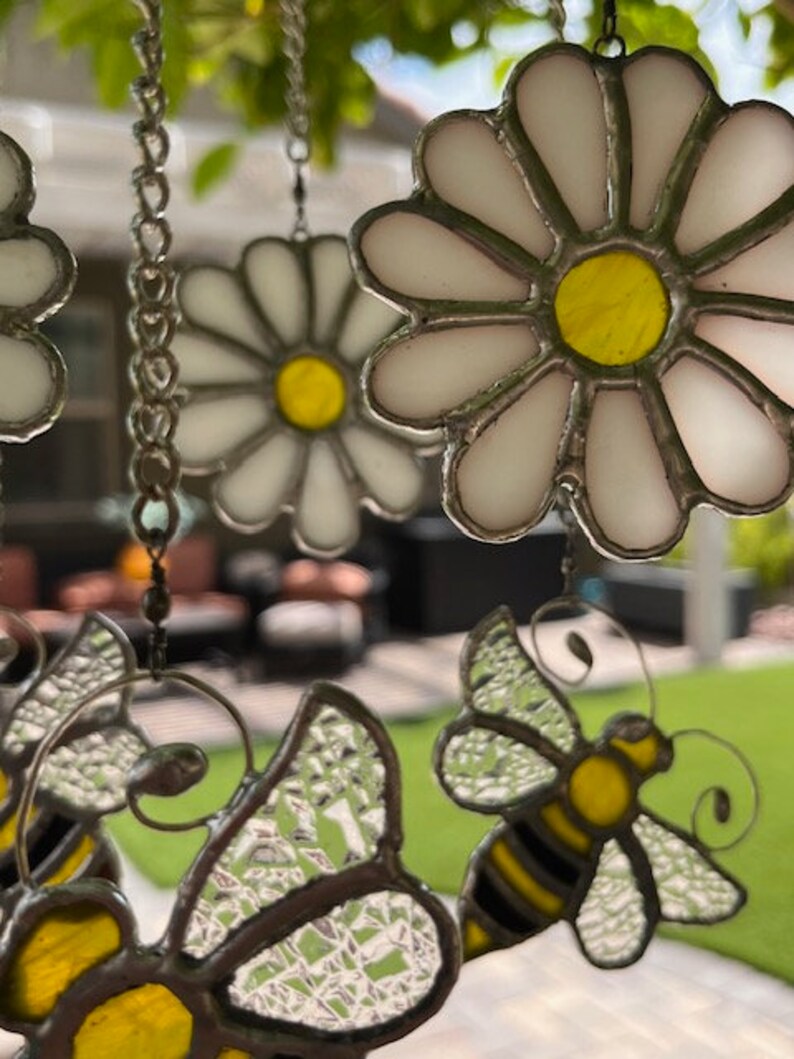 May include: A stained glass wind chime featuring white daisies with yellow centres and clear glass bumblebees with black stripes. The wind chime is hanging from a silver chain.