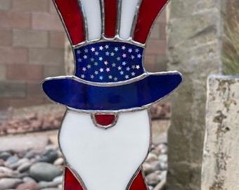 May include: A stained glass Uncle Sam decoration. The patriotic design features a red, white, and blue color scheme. The hat has red and white stripes, a blue band with stars, and a white beard. It hangs from a chain.