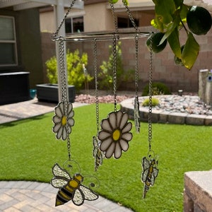 May include: A stained glass mobile featuring a white daisy, a bee, and a butterfly design. The mobile is suspended from a chain and hangs in the air.